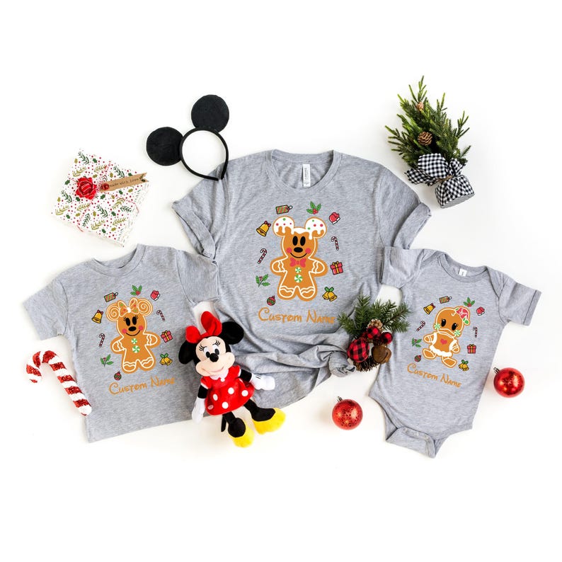 May include: A set of three grey t-shirts featuring a gingerbread Mickey Mouse design. The shirts are available in adult and child sizes. The shirts have the text "Custom Name" printed on them.