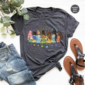 May include: A heather dark grey t-shirt with a graphic of seven princesses standing back to back with their hair up. The princesses are wearing different colored dresses and are surrounded by stars.