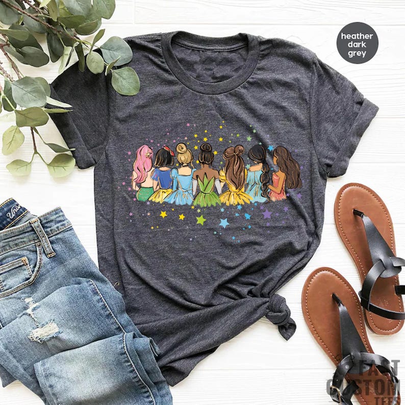 May include: A heather dark grey t-shirt with a graphic of seven princesses standing back to back with their hair up. The princesses are wearing different colored dresses and are surrounded by stars.