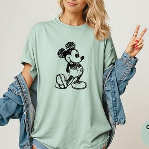 May include: A light green t-shirt featuring a black sketch of Mickey Mouse. The shirt is a Comfort Colors Bay, with a relaxed fit. The design is a classic cartoon character.