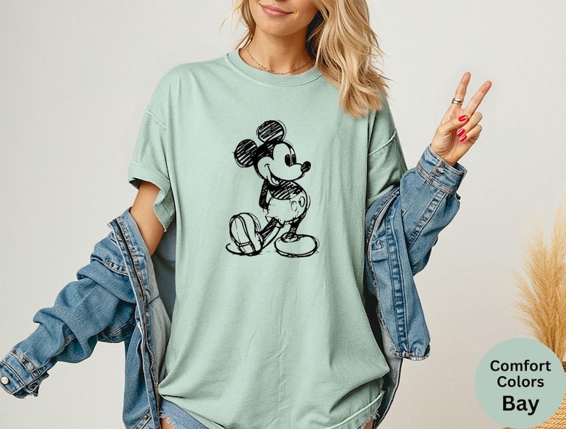 May include: A light green t-shirt featuring a black sketch of Mickey Mouse. The shirt is a Comfort Colors Bay, with a relaxed fit. The design is a classic cartoon character.