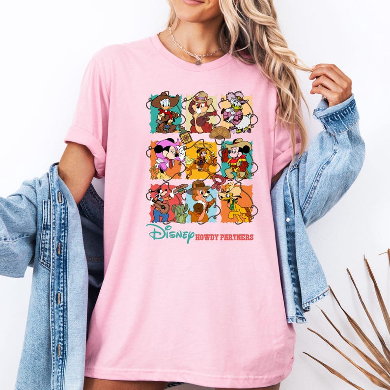 May include: A light pink t-shirt featuring a colorful graphic of Disney characters in cowboy attire. The design includes Mickey Mouse, Donald Duck, and other characters. The text "Disney Howdy Partners" is printed below the graphic.