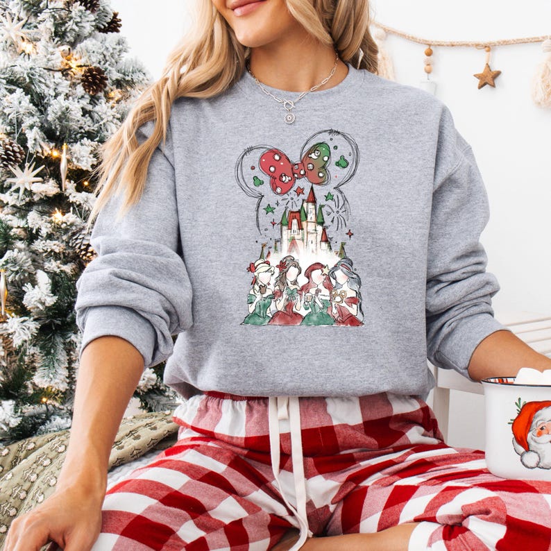May include: A gray sweatshirt featuring a festive design. The graphic includes a castle, fireworks, and a bow-shaped design with red and green accents. Below the castle are illustrations of characters in holiday attire. The sweatshirt is paired with red and white plaid pajama bottoms.