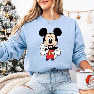 May include: A pale blue sweatshirt with a Mickey Mouse graphic. The cartoon character has black ears, a smiling face, white gloves, red shorts, and yellow shoes. The design is centered on the front of the long-sleeved top.