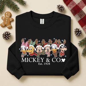 K&ouml;nnte beinhalten: Schwarzes Sweatshirt mit Disney-Figuren als Rentiere mit Geweihen und Schals. Mickey, Minnie, Donald, Daisy, Goofy und Pluto sind abgebildet. Der Text "Mickey & Co. Est. 1928" ist darunter gedruckt.