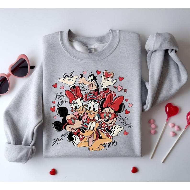 May include: A light gray sweatshirt featuring a graphic of Disney characters, including Mickey and Minnie Mouse, Donald and Daisy Duck, Goofy, and Pluto, surrounded by red hearts. The characters are wearing heart-shaped sunglasses and are surrounded by the words "Mickey Mouse", "Daisy Duck", "Minnie Mouse", and "Pluto".
