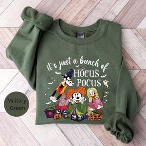 Op de afbeelding: Een groen sweatshirt met een afbeelding van Mickey Mouse, Goofy en Donald Duck verkleed als de Sanderson-zusters uit de film Hocus Pocus. De tekst "It's just a bunch of Hocus Pocus" staat op het sweatshirt gedrukt.