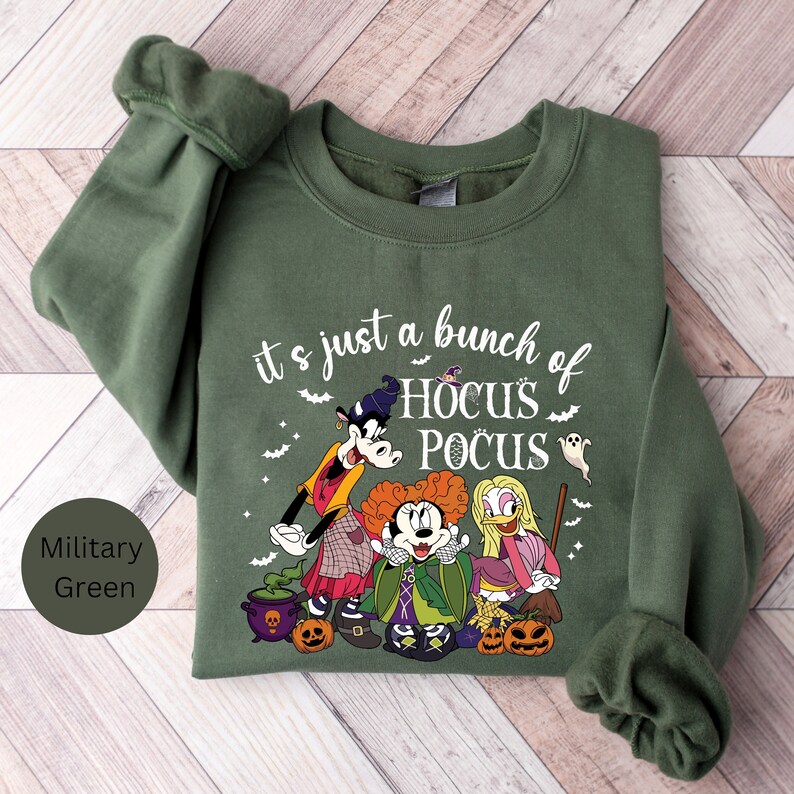 Op de afbeelding: Een groen sweatshirt met een afbeelding van Mickey Mouse, Goofy en Donald Duck verkleed als de Sanderson-zusters uit de film Hocus Pocus. De tekst "It's just a bunch of Hocus Pocus" staat op het sweatshirt gedrukt.