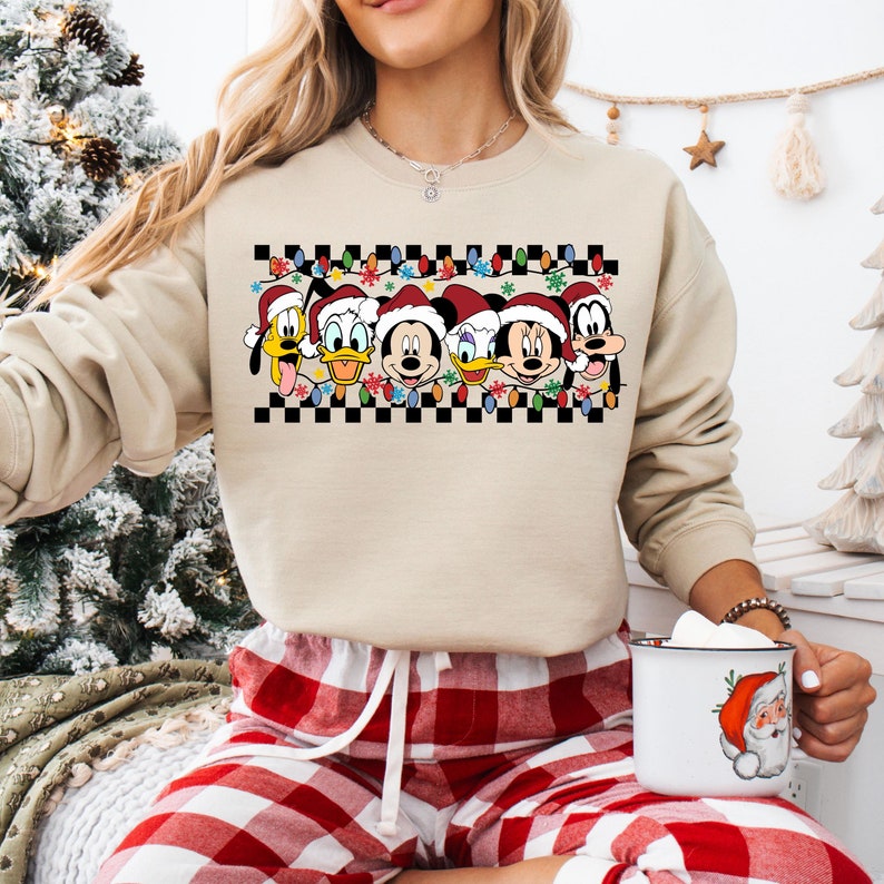 May include: A beige sweatshirt with a checkered pattern and a row of Disney characters wearing Santa hats. The characters include Mickey Mouse, Minnie Mouse, Donald Duck, Goofy, and Pluto. The characters are surrounded by Christmas lights.