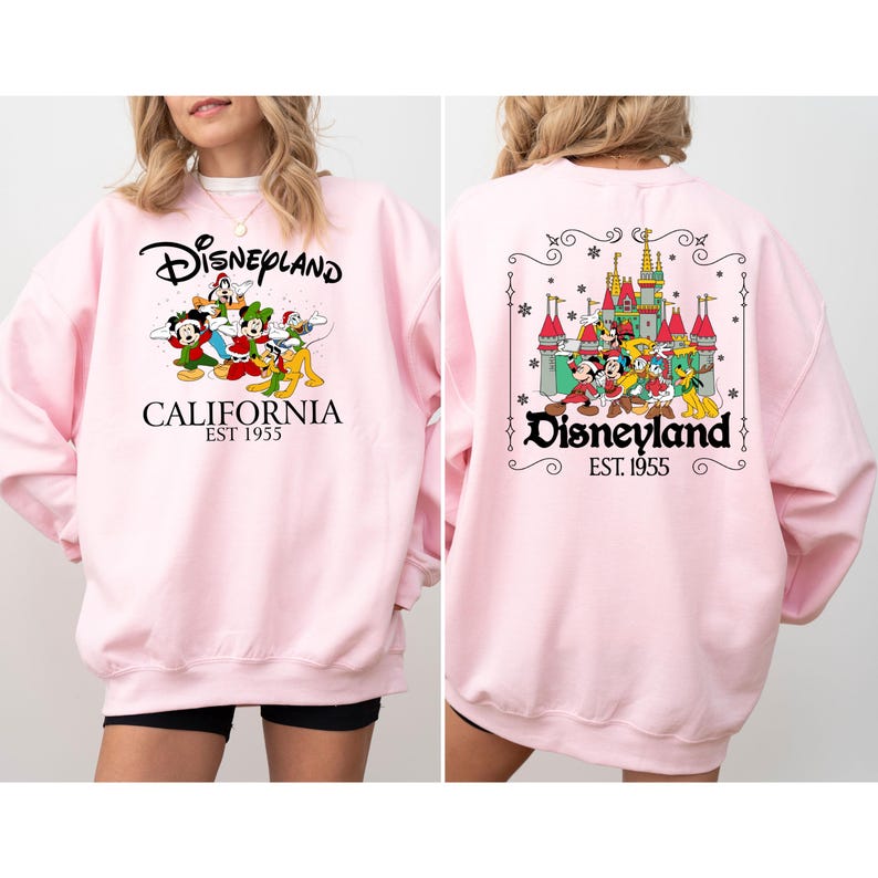 May include: A light pink sweatshirt featuring the Disneyland logo and various cartoon characters. The front of the sweatshirt displays the Disneyland logo with the text "California Est 1955". The back of the sweatshirt features a castle and more characters.