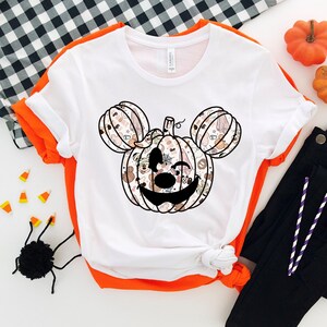 Op de afbeelding: Wit T-shirt met een zwart-wit Mickey Mouse pompoenontwerp. De pompoen is gemaakt van verschillende Halloween-afbeeldingen.