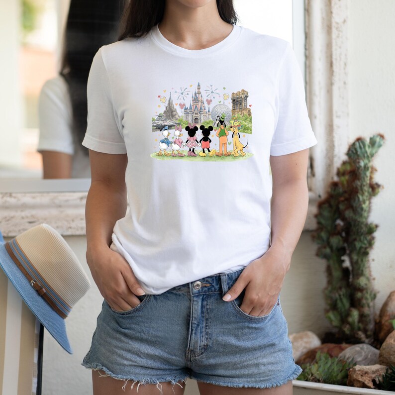 May include: White t-shirt featuring a colorful illustration of Disney characters in front of a castle and other landmarks. The graphic includes Mickey and Minnie Mouse, Donald and Daisy Duck, Goofy, and Pluto. The shirt is a classic crew neck style.