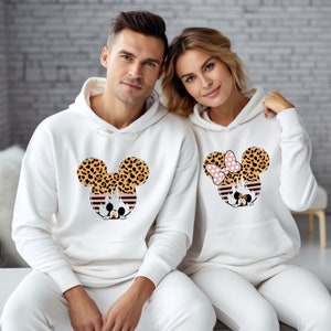 May include: A white hoodie with a leopard print Mickey Mouse design. The hoodie has a drawstring hood and a kangaroo pocket. The design features a pink bow on Minnie Mouse's head.