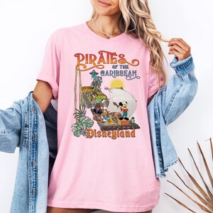 May include: Pink t-shirt with a vintage-style graphic of Mickey Mouse and other Disney characters on a pirate ship. The text reads "Pirates of the Caribbean Disneyland".
