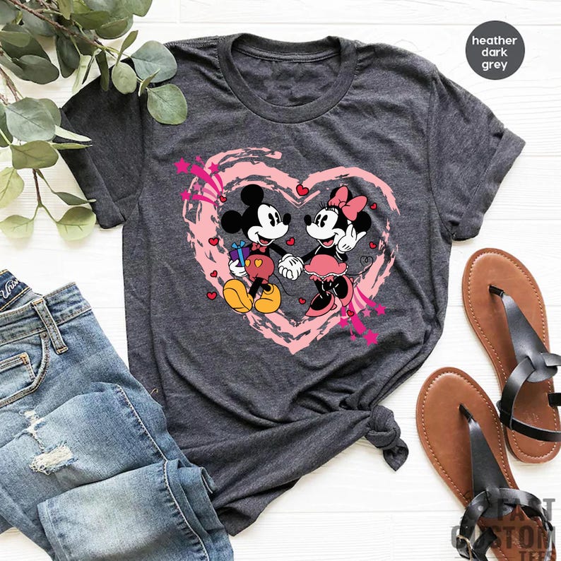 May include: Dark heather grey t-shirt featuring Mickey and Minnie Mouse holding hands inside a pink heart. Mickey Mouse is holding a gift. The shirt has a crew neck and short sleeves. The words "heather dark grey" are in the upper right corner.
