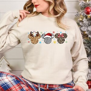 May include: A cream-colored sweatshirt with a Christmas-themed design featuring three Mickey Mouse ears with different animal print patterns. The ears are decorated with snowflakes, lights, and a Santa hat.