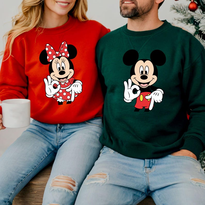 May include: Red sweatshirt featuring Minnie Mouse with a polka dot dress and bow. The sweatshirt is paired with light blue jeans. A person is holding a white mug. Another person wears a green sweatshirt with Mickey Mouse.