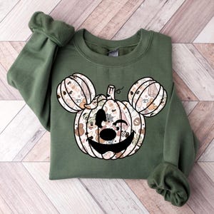 Op de afbeelding: Een groen sweatshirt met een pompoen-vormig Mickey Mouse hoofd ontwerp. De pompoen is gevuld met verschillende Halloween-thema patronen en personages.