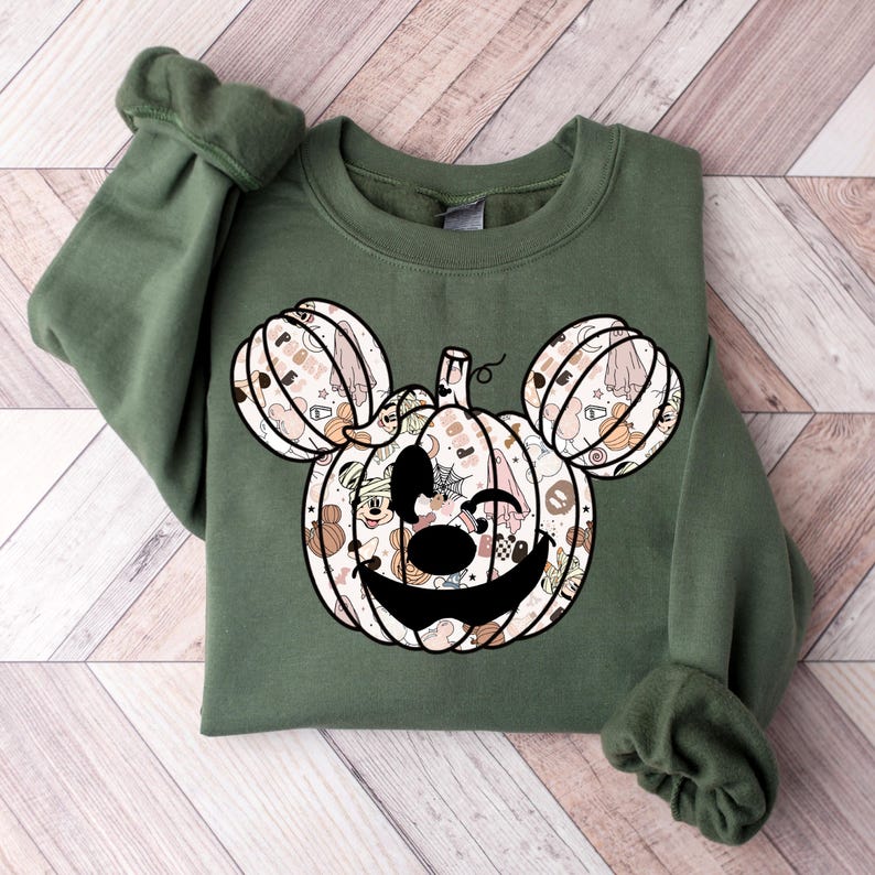 Op de afbeelding: Een groen sweatshirt met een pompoen-vormig Mickey Mouse hoofd ontwerp. De pompoen is gevuld met verschillende Halloween-thema patronen en personages.
