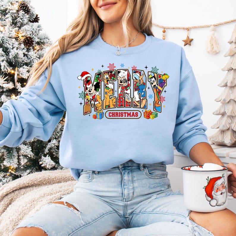 May include: A light blue sweatshirt with the words "MERRY CHRISTMAS" in a colorful, cartoonish font. The letters are filled with various cartoon characters. Presents and snowflakes surround the text. The person is holding a mug with a Santa Claus design.
