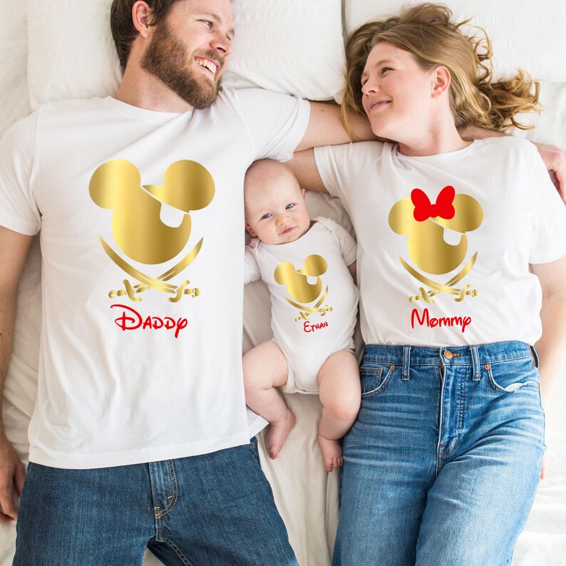 May include: A family of three wearing white t-shirts with a gold Mickey Mouse head design. The shirts say "Daddy", "Mommy", and "Ethan".
