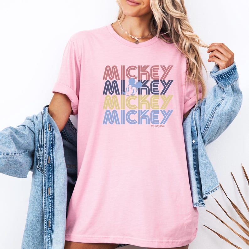 May include: A light pink t-shirt with the word "MICKEY" stacked four times in different colors. A Mickey Mouse silhouette is in the center. The text "THE ORIGINAL" is below the bottom "MICKEY".