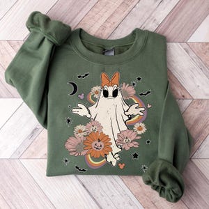 May include: A green sweatshirt with a white ghost wearing a pink bow and surrounded by flowers and stars. The ghost has a surprised expression and is holding its arms out. The sweatshirt has a rainbow design behind the ghost.