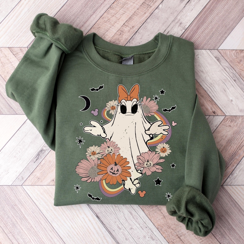 May include: A green sweatshirt with a white ghost wearing a pink bow and surrounded by flowers and stars. The ghost has a surprised expression and is holding its arms out. The sweatshirt has a rainbow design behind the ghost.