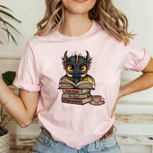 May include: A pink t-shirt with a cute cartoon dragon wearing glasses and reading a book. The text on the shirt reads "read books be kind stay weird".