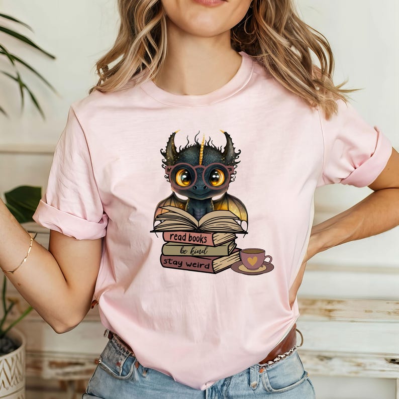 May include: A pink t-shirt with a cute cartoon dragon wearing glasses and reading a book. The text on the shirt reads "read books be kind stay weird".
