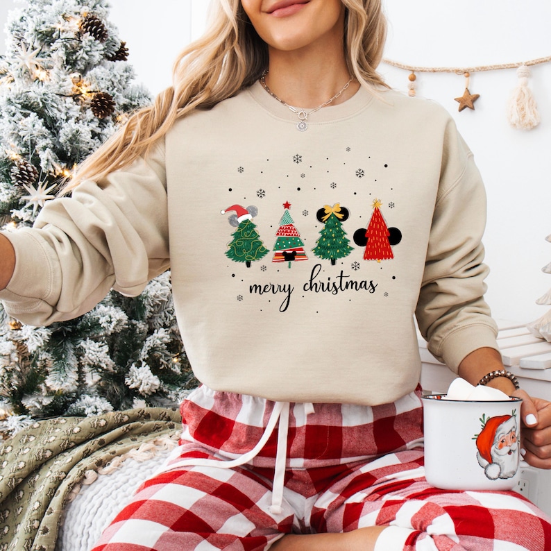 May include: Beige sweatshirt with a festive Christmas tree design. The graphic features four stylized Christmas trees with mouse ears and a Santa hat, with the words "merry christmas" below. The person is wearing red and white plaid pajama pants.