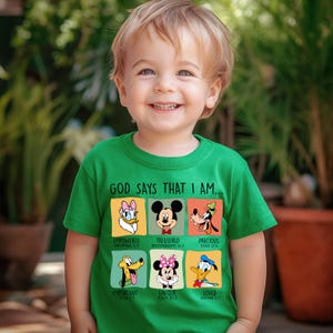 Puede incluir: Una camiseta verde con personajes de Disney y el texto "GOD SAYS THAT I AM...". La camiseta incluye im&aacute;genes de Daisy, Mickey Mouse, Goofy, Pluto, Minnie Mouse y Donald, con palabras como "Empowered" y "Loved".