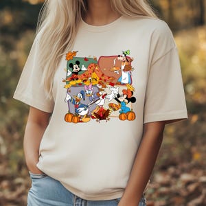 May include: Beige t-shirt featuring a colorful graphic of Disney characters celebrating Thanksgiving. Mickey Mouse, Minnie Mouse, Donald Duck, Daisy Duck, and Goofy are depicted with pumpkins and autumn leaves. The shirt has a crew neck and short sleeves.