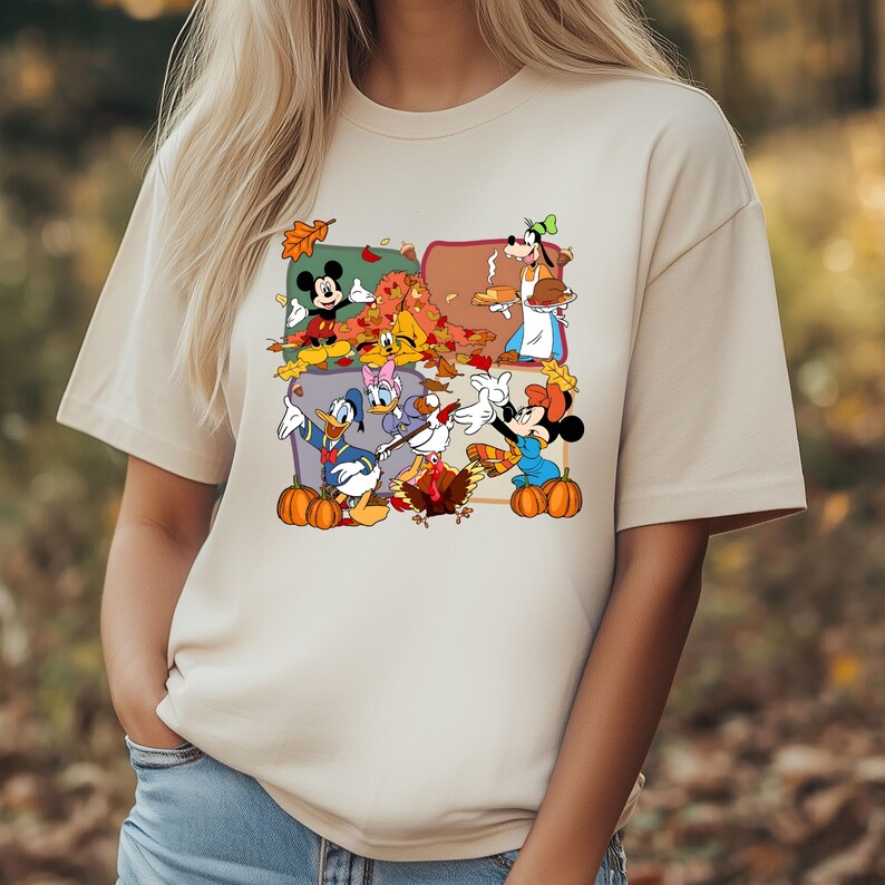May include: Beige t-shirt featuring a colorful graphic of Disney characters celebrating Thanksgiving. Mickey Mouse, Minnie Mouse, Donald Duck, Daisy Duck, and Goofy are depicted with pumpkins and autumn leaves. The shirt has a crew neck and short sleeves.