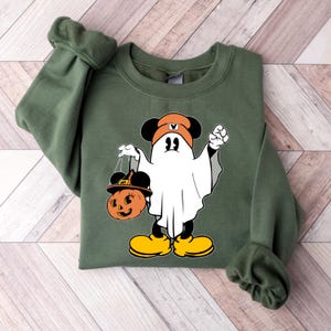May include: Olive green sweatshirt featuring a Halloween-themed graphic. The graphic depicts a cartoon ghost character with Mickey Mouse ears, wearing a hat and holding a pumpkin-shaped bag. The character is wearing yellow shoes. The sweatshirt is folded.