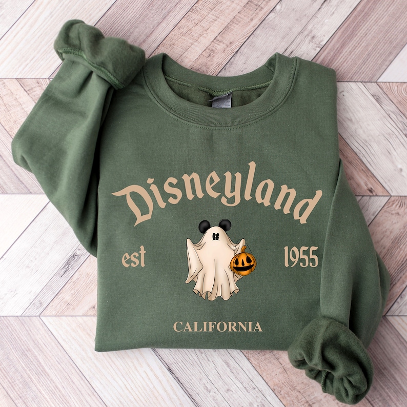 Puede incluir: Sudadera verde con un fantasma blanco que sostiene una calabaza. El texto "Disneyland est 1955 CALIFORNIA" est&aacute; impreso en la parte delantera.