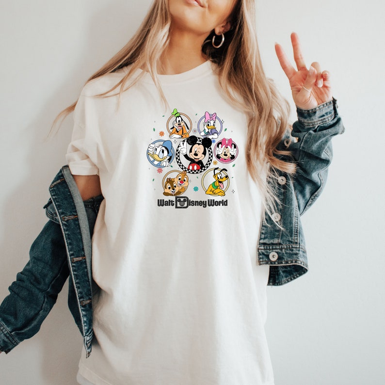 Op de afbeelding: Wit T-shirt met een kleurrijke afbeelding van Mickey Mouse en andere Disney-figuren in cirkels. De tekst "Walt Disney World" is onder de afbeelding gedrukt.