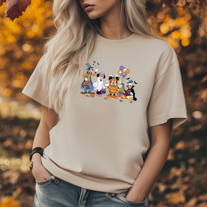 May include: Beige t-shirt featuring a Halloween-themed graphic of cartoon characters including Mickey Mouse, Minnie Mouse, Donald Duck, and Goofy dressed in costumes. The graphic includes pumpkins, ghosts, and balloons.