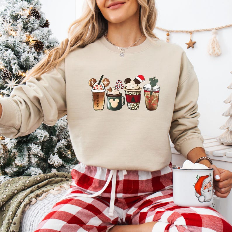May include: Beige sweatshirt featuring a festive graphic of four holiday-themed coffee drinks. Each drink has unique designs, including Mickey Mouse ears, a Santa hat, and a shamrock. The model is wearing red and white plaid pajama pants and holding a mug with a Santa Claus design.