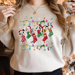 May include: A cream-colored sweatshirt with a Christmas-themed design featuring Mickey Mouse, Donald Duck, Goofy, and Pluto wearing Christmas stockings. The design includes a string of lights and snowflakes.