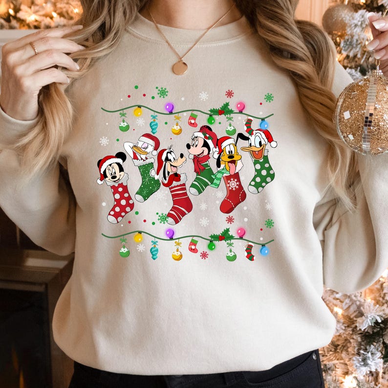 May include: A cream-colored sweatshirt with a Christmas-themed design featuring Mickey Mouse, Donald Duck, Goofy, and Pluto wearing Christmas stockings. The design includes a string of lights and snowflakes.