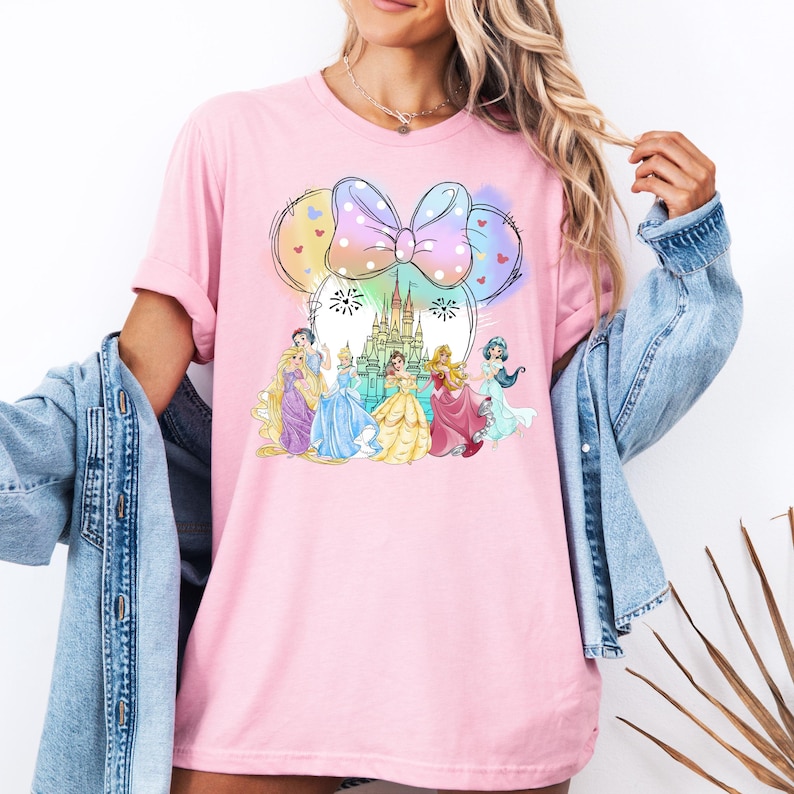 May include: Pink t-shirt with a graphic of Disney princesses in front of Cinderella's castle with a Minnie Mouse bow and balloons.