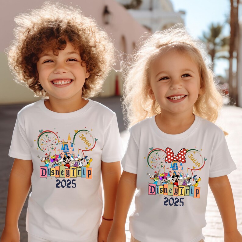 May include: Two children wearing white t-shirts with a colorful Disney Trip 2025 design. The shirts feature Mickey Mouse and other Disney characters, fireworks, and the words "Disney Trip 2025". One shirt has the name "John" and the other has the name "Julia".