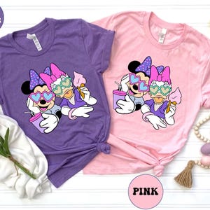 Pode incluir: Duas camisetas com uma estampa gr&aacute;fica de Minnie Mouse e Daisy Duck usando &oacute;culos de sol e segurando cones de sorvete. As camisetas s&atilde;o roxas e rosa com o texto "TEAM PURPLE" e "PINK" nelas.