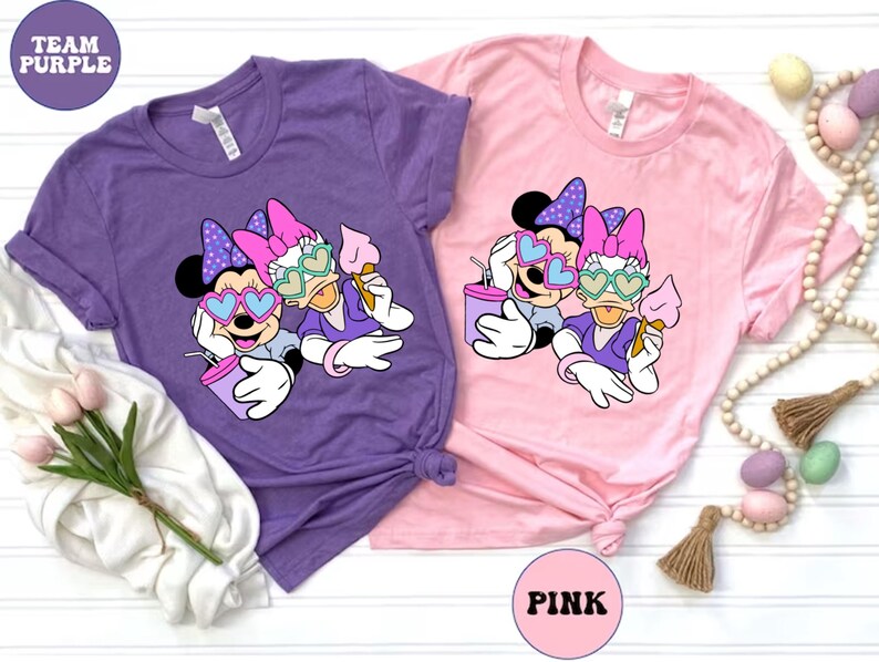 Pode incluir: Duas camisetas com uma estampa gr&aacute;fica de Minnie Mouse e Daisy Duck usando &oacute;culos de sol e segurando cones de sorvete. As camisetas s&atilde;o roxas e rosa com o texto "TEAM PURPLE" e "PINK" nelas.
