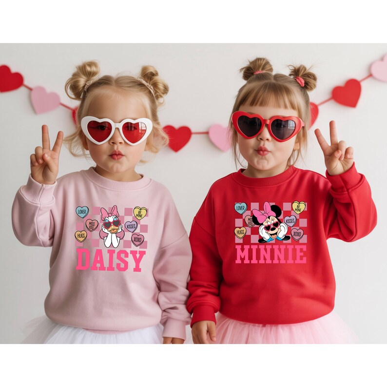 May include: Two young children wearing heart-shaped sunglasses and sweatshirts. One sweatshirt is light pink with a Daisy Duck graphic and the name "DAISY". The other is red with a Minnie Mouse graphic and the name "MINNIE". Both sweatshirts have candy heart graphics.
