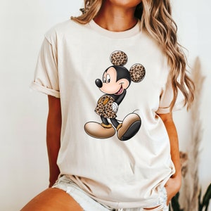 Op de afbeelding: Een beige T-shirt met een cartoon afbeelding van Mickey Mouse die een outfit met luipaardprint draagt. Mickey Mouse glimlacht en heeft zijn handen op zijn heupen.