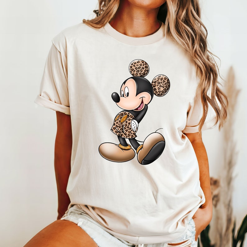 Op de afbeelding: Een beige T-shirt met een cartoon afbeelding van Mickey Mouse die een outfit met luipaardprint draagt. Mickey Mouse glimlacht en heeft zijn handen op zijn heupen.