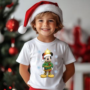 Pode incluir: Uma crian&ccedil;a vestindo uma camiseta branca com Mickey Mouse em uma roupa de Natal, um gorro de Papai Noel e flocos de neve. O personagem de desenho animado est&aacute; vestido com um su&eacute;ter verde e sapatos amarelos. A crian&ccedil;a tamb&eacute;m est&aacute; usando um gorro de Papai Noel vermelho e branco.