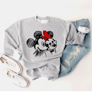 May include: A light gray sweatshirt featuring a black and red graphic of Mickey and Minnie Mouse. The sweatshirt is paired with light blue jeans, white sneakers, and a white background. The graphic design is a sketch-like illustration.